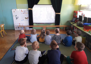 Dzieci siedzą na dywanie i oglądają prezentację multimedialną.