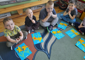 Dzieci siedzą na dywanie i prezentują swoje prace.
