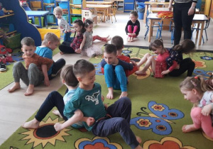 Dzieci siedzą na dywanie dotykając sie plecami i przepychają się.