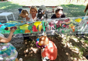 Dzieci i panie maluja farbami łąkę.