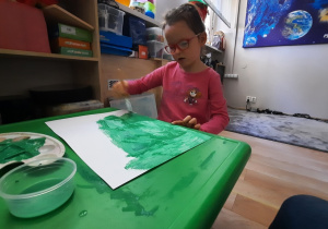 Dziewczynka zamalowywuje zieloną farbą kartk papieru.