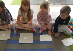 Dzieci siedzą na dywanie i słuchają instrukcji wykonania pracy.