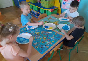 Dzieci siedzą przy stole i malują talerzyki papierowe farbami.