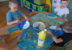 Chłopcy siedzą przy stole i malują talerzyki papierowe farbami.