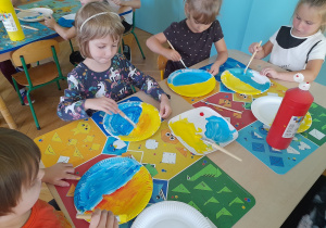 Dzieci siedzą przy stole i malują talerzyki papierowe farbami.