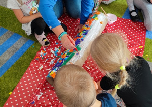 Dzieci na dywanie oklejają kawałkami papieru kolorowego tubę.