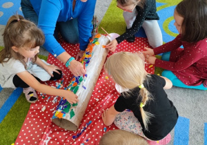 Dzieci na dywanie oklejają kawałkami papieru kolorowego tubę.