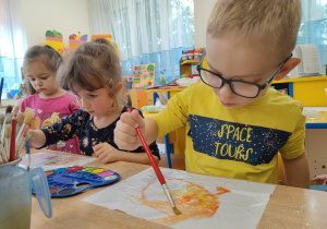 Dzieci malują pędzlaem po kartce jesienne krajobrazy.
