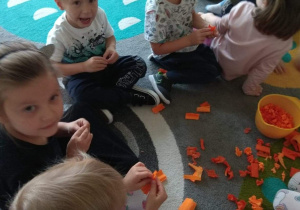 Dzieci siedzą na dywanie i bawią się pomarańczową bibułą.