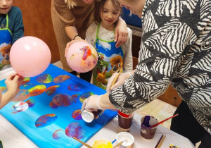 Dziewczynka wspólnie z artystką odciska balony umoczone farbą na płótnie.
