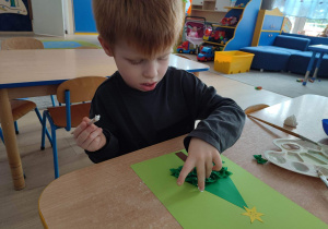 Chłopiec przykleja zielony makaron kokardka do kartki.