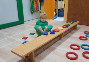 Chłopiec siedzi na podłodze i stopami układa woreczki na ławeczce.