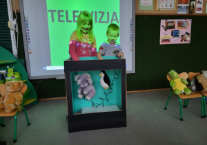Dzieci animują maskotkami w telewizorze wykonanym z pudła kartonowego.