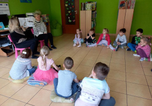 Dzieci siedzą na podłodze, słuchają czytanej książki.