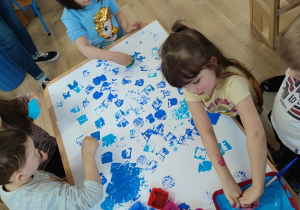 Dzieci tapują foremkami białe tło niebieską farbą.