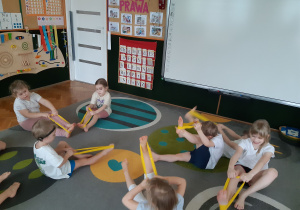 Dzieci siedzą na dywanie i ćwiczą z gumowymi szarfami.