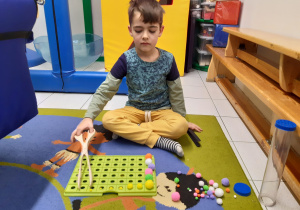Chłopiec siedzi na dywanie i szczypcami układa kolorowe pompony.
