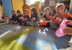 Dzieci sidza na dywanie, w dłoniach trzymają kubeczki i wystukują rytm podany przez nauczycielkę.