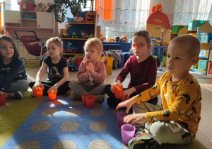 Dzieci z plastykowymi kubeczkami siedzą na dywanie.