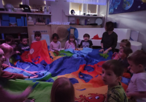 Dzieci siedzą na podłodze przykryte chustą animacyjną, w sali są zgaszone światła.