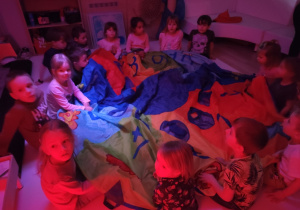 Dzieci siedzą na podłodze przykryte chustą, obserwują kolejne włączanie kolorowych urządzeń.