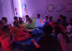 Dzieci siedzą na podłodze przykryte chustą, obserwują kolejne włączanie kolorowych urządzeń.