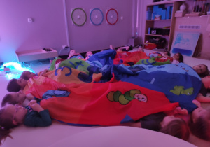 Dzieci leżą na podłodze przykryte chustą, obserwują kolejne włączanie kolorowych urządzeń.