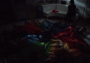 Dzieci leżą na podłodze przykryte chustą w całkowitej ciemności.