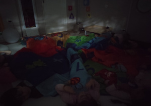 Dzieci leżą na podłodze przykryte chustą w całkowitej ciemności.