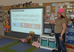 Nauczycielka z maską świnki odczytuje zagadkę z tablicy multimedialnej.