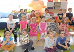 Dzieci siedzą na krzesłach i dywanie i słuchają opowieści o pszczołach.
