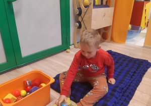 Chłopiec siedzi na materacu piankowym i chwyta stopami piłkę.