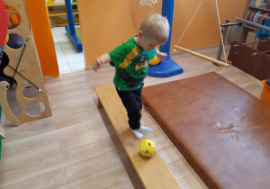 Chłopiec idzie i turla piłkę po ławeczce.