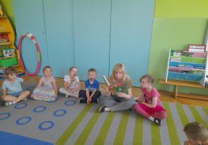 Dzieci siedzą na dywanie i słuchają książki czytanej przez nauczycielkę.