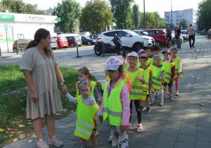 Dzieci z nauczycielką spacerują w parach po chodniku.