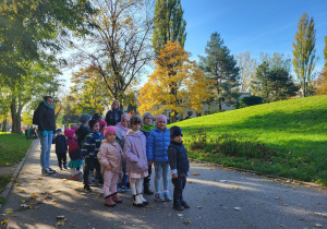 Dzieci pozują do zdjęcia na tle budynku przedszkola, jesiennego drzewa i błękitnego nieba.