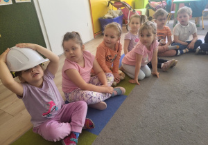 Dzieci siedzą na dywanie, jedna dziewczynka przymierza kask ochronny.