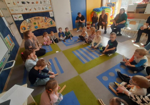 Dzieci w kole siedzą trzymając patyki w dłoni.