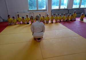 Dzieci siedzą na macie w szeregu, przed nimi trener judo.