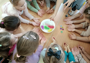 Dzieci siedza przy stole z wyciągniętymi rękoma, na stole jest talerzyk z brokatem.