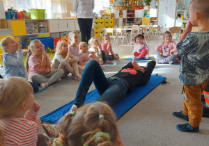 Pani leży na macie, dzieci obserwują.