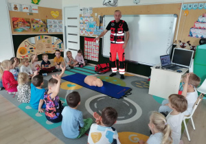 Dzieci siedzą i słuchają ratownika.Chłopiec uciska manekina.