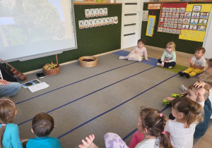 Dzieci siedzą na dywanie, oglądają zdjęcia na ekranie.