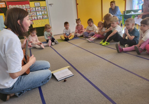 Pani gran na instrumencie, dzieci siedzą na dywanie.