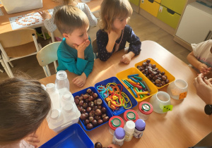 Dzieci siedzą przy stoliku, na którym leżą kasztany, plastelina i inne akcesoria.