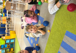 Dzieci leżą na brzuchu, na dywanie z piłeczkami - wieksza i mniejszą.