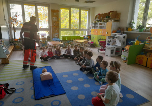 Dzieci siedzą na dywanie i słuchają Pana ratownika.