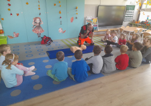 Dzieci siedzą na dywanie i słuchają Pana ratownika.
