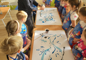 Pani rozlewa farbę na kartonach, dzieci obserwują.