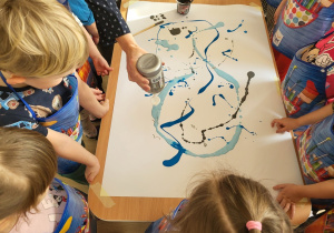 Pani rozlewa farbę na kartonach, dzieci obserwują.